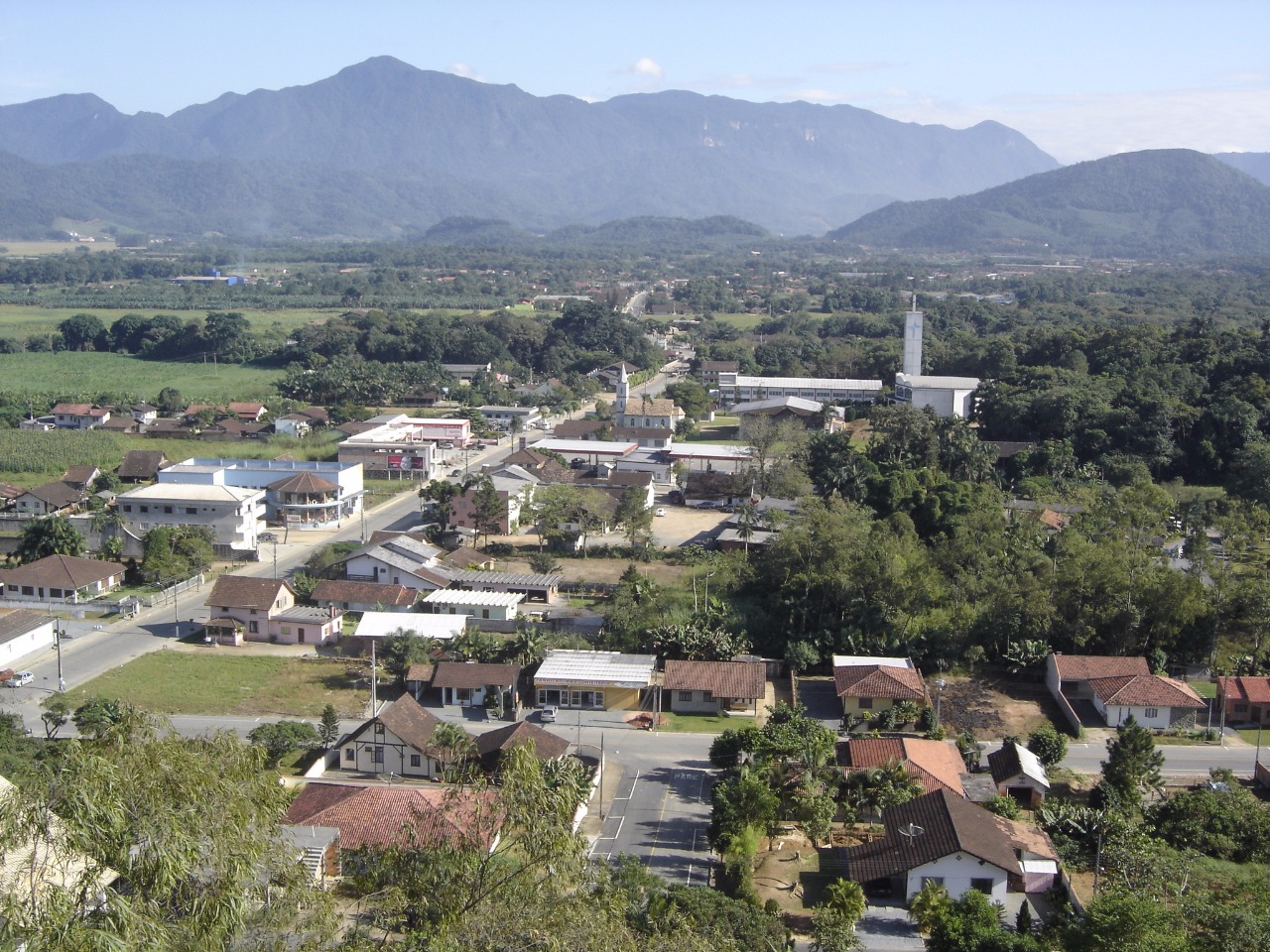 Vista aérea de Schroeder. A imagem mostra montanhas, céu azul, uma rua e várias construções rodeadas pela natureza conservada há mais de 56 anos.